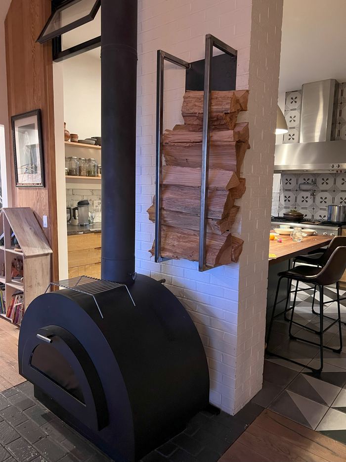 Raw steel firewood rack beside a wood stove, loaded with split wood