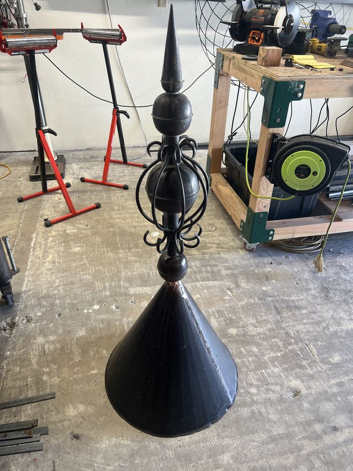 Decorative steel spire with a ball, scrollwork, and a cone base, standing in the shop